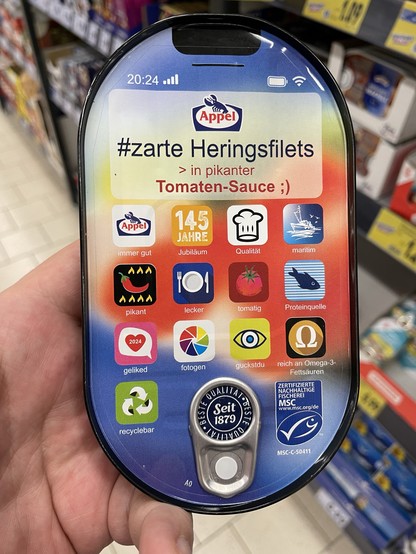 Man sieht eine in der Hand gehaltene Dose mit Fischfilets. Die Front der Dose ist wie eine Handyoberfläche gestaltet, mit vollem Empfang und eingeschaltetem WLAN. Einige Logos und andere Grafiken sind als Icon gestaltet, darunter steht z.B. „pikant“, „lecker“ oder „tomatig“. Dort wo eventuell ein Datumswidget wäre im oberen Drittel der Dose steht ein dreiteiliger Text:

#zarte Heringsfilets
> in pikanter
Tomaten-Sauce ;)
