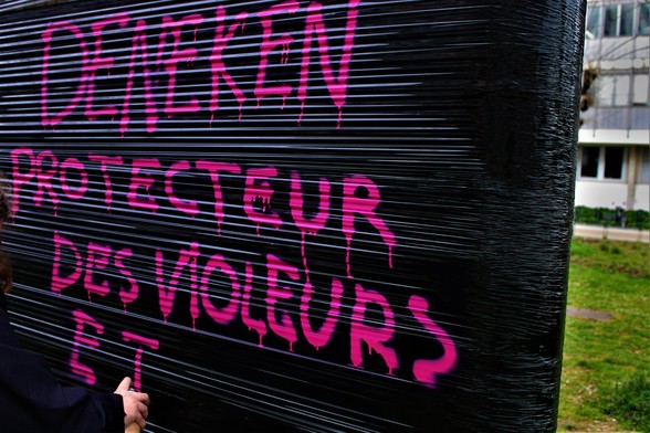 Un•e bloqueur•euse en train de taguer "Deneken protecteur des violeurs" sur une bande de plastique noir accrochée entre deux arbres