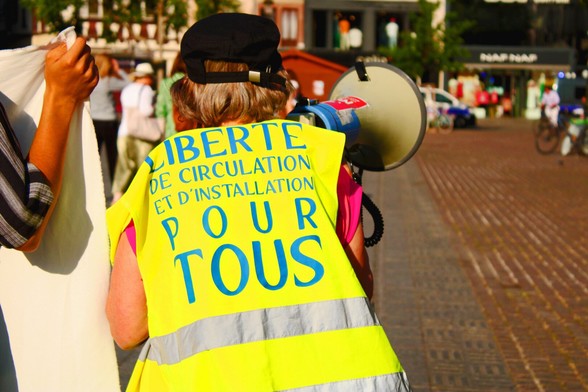 Un•e manifestant•e avec un mégaphone portant l'inscription "Liberté de circulation et d'installation pour tou•tes"