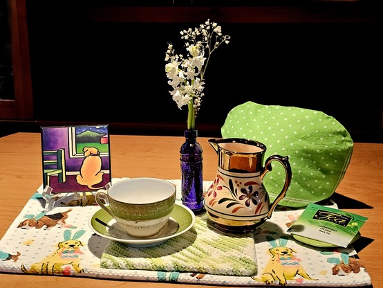 Sunlit tea tableau in bright green and calm purple with a painting of a dog seen from behind, looking out a window as if watching for something. The tea towel features dogs dressed in bunny ears with Easter baskets hanging from their mouths. There is a very old cup and saucer set and a 1930s milk pitcher, and a vintage purple glass mini bottle holding white Spanish Bluebell blossoms. The tea is Strawberry Lychee black tea from Hawaiian Islands Tea Company.