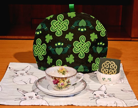 Primarily green tea tableau with Celtic knotwork and claddagh motif on tea cozy and tea packet of Irish Breakfast black tea, on a tea towel decorated with quizzical looking rabbits, with an antique cup and coordinating vintage saucer, and a small bowl decorated with a shamrock.