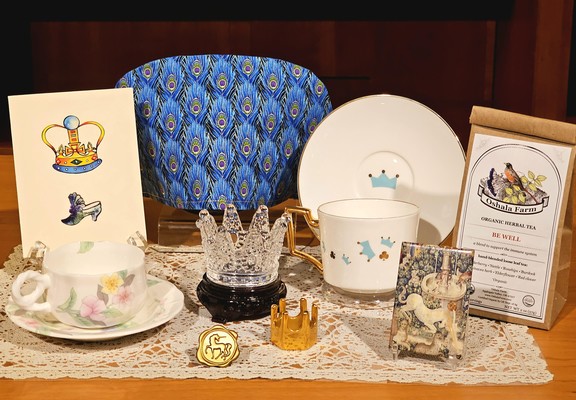 Tea tableau with a peacock feather pattern tea cozy, a crystal crown, a gold crown, a gold medallion showing a unicorn, a small picture of a Bayeux Tapestry detail of a unicorn, a card with a crown and fancy slippers, vintage china, and  herbal wellness tea from Oregon.