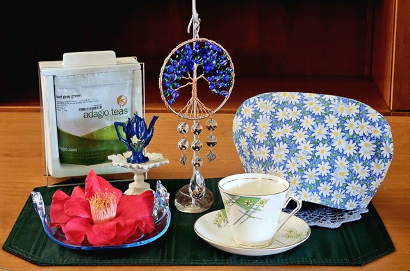 Tea tableau centering around a green and blue jewel beaded Tree of Life suspended on a display stand. The tea cozy is field of daisies, the vintage cup and saucer have a daisy design, and there is a large dark red Japanese Camellia blossom on an aquarium blue Depression Glass dish. There is a miniature dark blue teapot on a scalloped pedestal, and the tea is Earl Grey Green tea from Adagio.