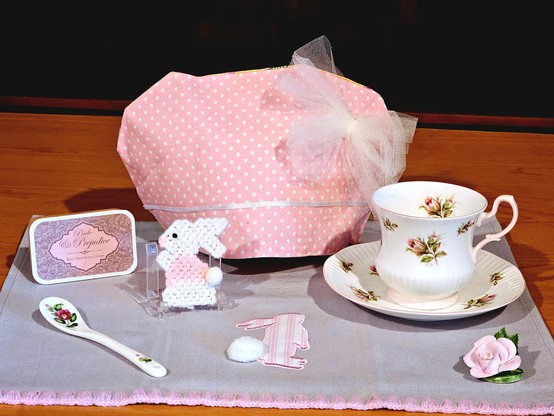 Tea tableau in pink and grey. There is a pink polkadotted cozy with a frothy white bow, a vintage rosebud patterned cup and saucer with a graceful pink handle, an embroidered pink striped bunny, a needlework pink and white bunny, a porcelain rose, a rose patterned ceramic spoon, and a pink tin of customer blended black tea flavored with rose, strawberry, and cream from Adagio, called Pride and Prejudice, all on a grey tea towel with pink trim.