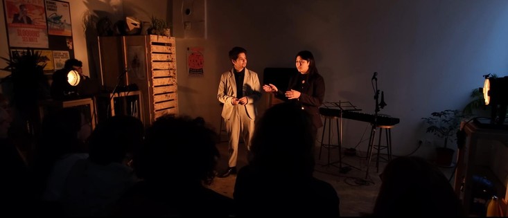 Image en format très large, cinéma. Un drag king blanc en costume beige et chemise noire, cheveux courts et bouc, et une personne métis franco-chinoise aux cheveux longs et foncés, dans une lumière très chaleureuse arrivant sur elleux depuis les côtés. Sur le mur blanc au fond, à droite contraste la lumière du jour qui paraît très bleue. Des affiches et un peu de mobilier en bois sur la gauche. À l'avant-plan, des silhouettes du public. Salle d'environ 7m de large et haute de plafond (on ne le voit pas).