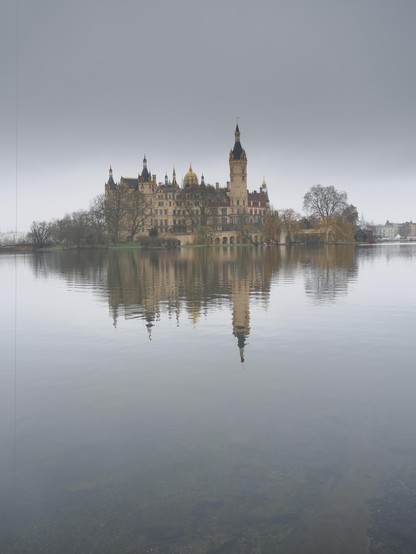 The image depicts a large, historic castle with numerous spires and towers, situated on a piece of land surrounded by a body of water. The castle is reflected in the calm water, and there are trees and muted vegetation around it, under a cloudy sky.