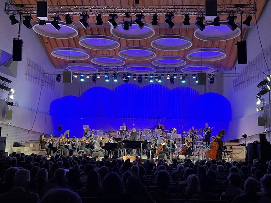Pippo Pollina am Klavier in der Mitte umgeben vom Jugensymphonieorchester vor blauem Hintergrund.