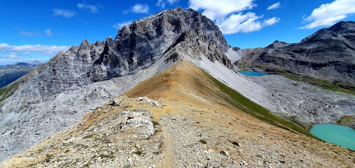 Foto einer Berglandschaft. Im Vordergrund ein Grat aus gelb-orangem Gestein, dahinter ein Gipfel und rechts zwei Bergseen.