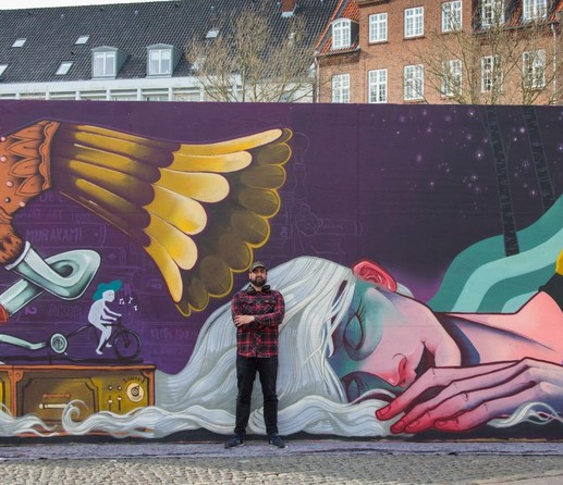 Mural by Malakkai of woman sleeping on her right side. Her white hair spills over her. A quirky phonograph record player looms over her, as if in dream. The artist stands in front of the mural.