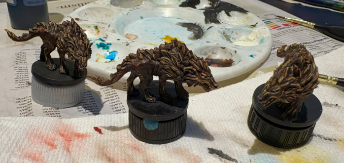Three miniatures of wolves on risers at a painting station. They are painted brown with light fur highlights and yellow eyes. A well-used palette is in the background.