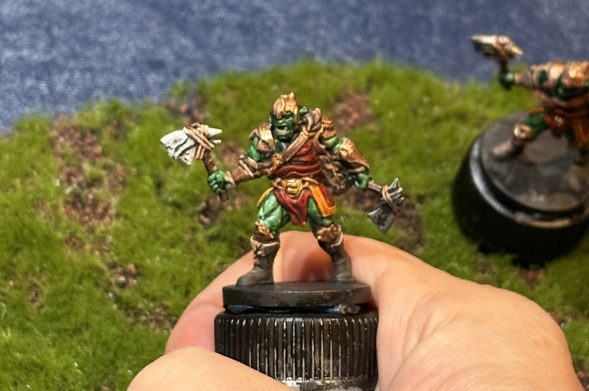 Close-up of a hand holding a painted miniature of an orc. The orc has green skin, a red and yellow tunic, bronze armor, and an axe in each hand.  Another similar figure is seen, blurry, in the background of what looks like green grass and blue plastic.