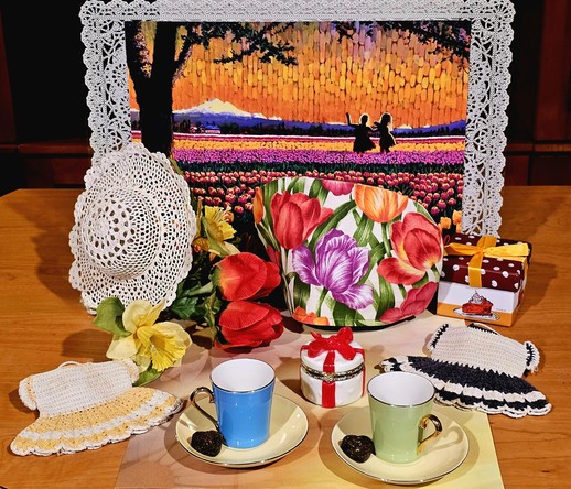 Tea tableau in front of a vibrant watercolor painting of two little girls in silhouette dancing in a field of tulips with Mt Baker in the background. The tea cozy is a tulip print, there are a lace hat and two crocheted doll dresses, a ceramic trinket box shaped like a present tied with a red bow, a gift box decorated with a birthday cake, a bouquet of tulips and daffodils, two vintage demitasse sets in sherbet colors, and two heart shapes of pressed tea leaves.