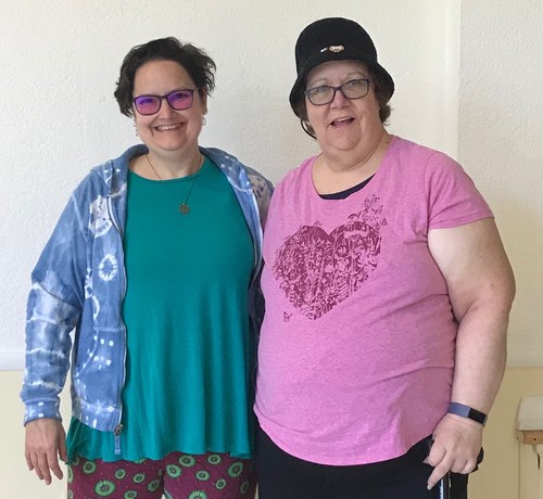 Two larger bodied white women with short dark hair and glasses. I am wearing a green top with an indigo tie-dyed hoodie and red leggings with green circles. I am standing with Vicki who has a pink shirt with a heart design in red and a black hat, she also has a cane in her left hand.