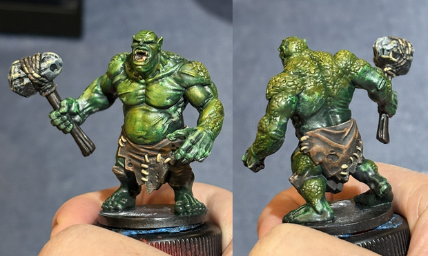 Front and back view of a hand holding a wargaming miniature of an ogre.  It has been painted a greenish color with rough patches of his skin fading to a yellowish green.  He's wearing a leather skirt and holding a big stone hammer.