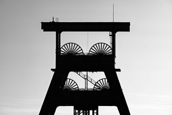 A black-and-white silhouette of a historic mining tower with large pulley wheels, set against a pale sky. The architectural structure is sharply defined, showcasing industrial symmetry and legacy.