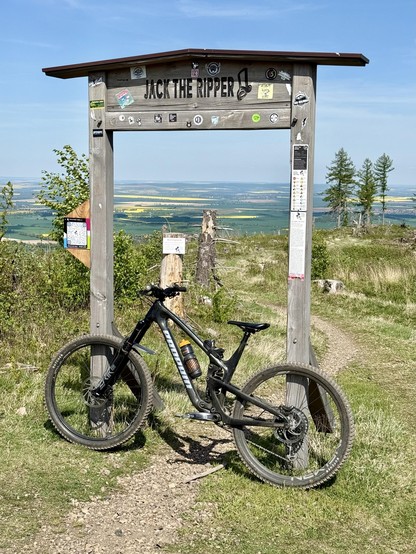 Ein Mountainbike lehnt sich an einen hölzernen Tor mit der Aufschrift "JACK THE RIPPER", umgeben von Aufklebern. Der Hintergrund bietet einen malerischen Blick auf Hügel und Felder unter einem klaren blauen Himmel.