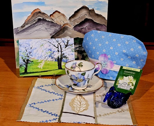 Springtime tea tableau with a watercolor mountainscape in the background, a small oil painting of a cherry grove in full blossom,  a light blue calico tea cozy embellished with a pink silk flower and white ribbon, vintage white and blue floral china, a cobalt glass swan, all on a piecework embroidered mat in white with blue and golden thread. The tea is Jasmine green tea from Ahmad.