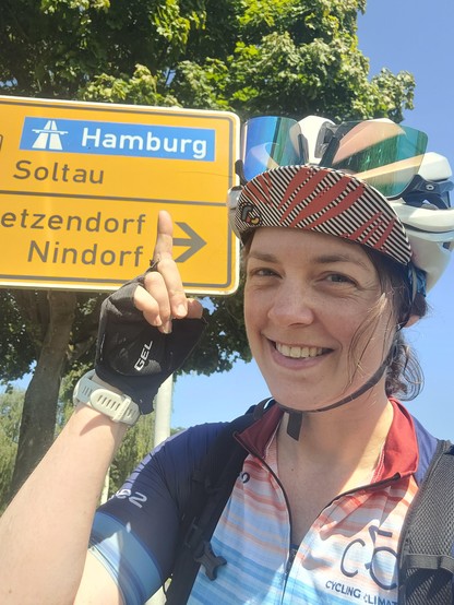Selfie, mon visage souriant, portant le maillot de la cyclo pour le Climat, gapette et casque. Pointant du doigt un panneau indiquant Hamburg derrière moi