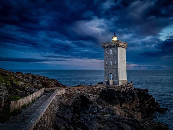 Rechteckiger Leuchtturm kurz nach Sonnenuntergang, umgeben von Felsen und Meer, unter bewölktem Himmel.