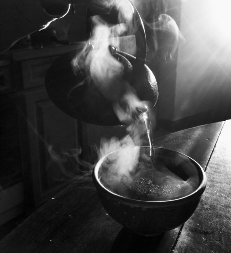A hand pours hot tea from a cast-iron teapot into a stoneware bowl. The rising sun's low-angled rays enhance the curls of steam. (Version françæ) Æn main verse dal thé brûlanx d’æn théière en fonte dans æn bol en grès. Læs volutes de vapeur sont miz en valeur par les rayons rasanz dal soleil levanx.