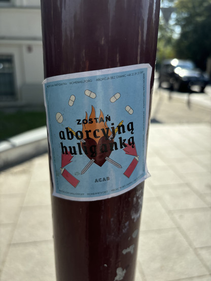 Sticker depicting a burning heart that is pierced with two crossed swords. There are two spray cans and pills around it. On top, there is a text: Zostań aborcyjna huliganką. ACAB. aborcyjna partyzantka, womenhelp.org, aborcja bez granic +48 22 29 597