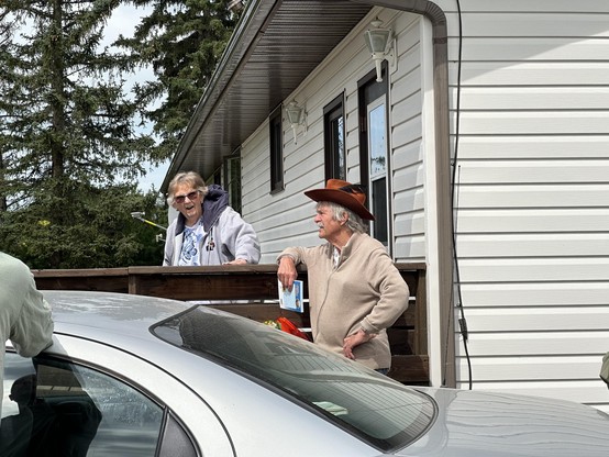 Two people outside of a house. There is an older woman, Marion Knudsen along with an older man, Tako, who is leading the field trip. There is a car in the foreground.