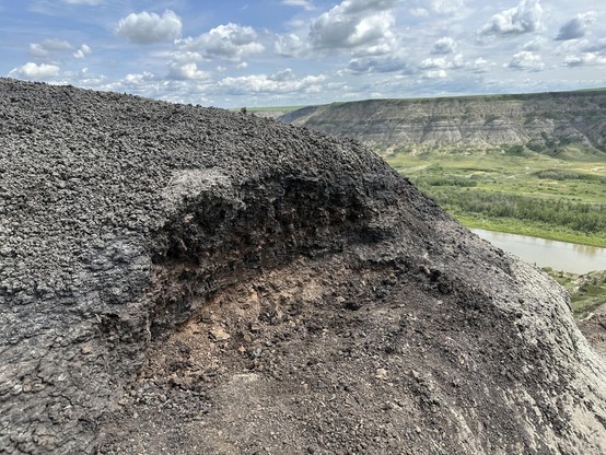 The side of a hill with a small excavation. In the excavation you can see the colour change from brown / black to orange; that's the K-Pg extinction layer. In the background you can see the badlands and the river running through them.