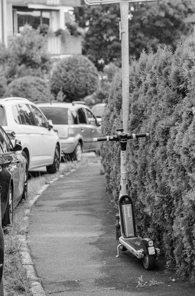 Das Bild zeigt eine Schwarzweißaufnahme einer Straßenszene. Ein E-Scooter steht neben einem dichten Busch auf dem Bürgersteig. Im Hintergrund sind geparkte Autos und Gebäude zu sehen. Das Foto ist von einer etwas erhöhten Position aufgenommen.