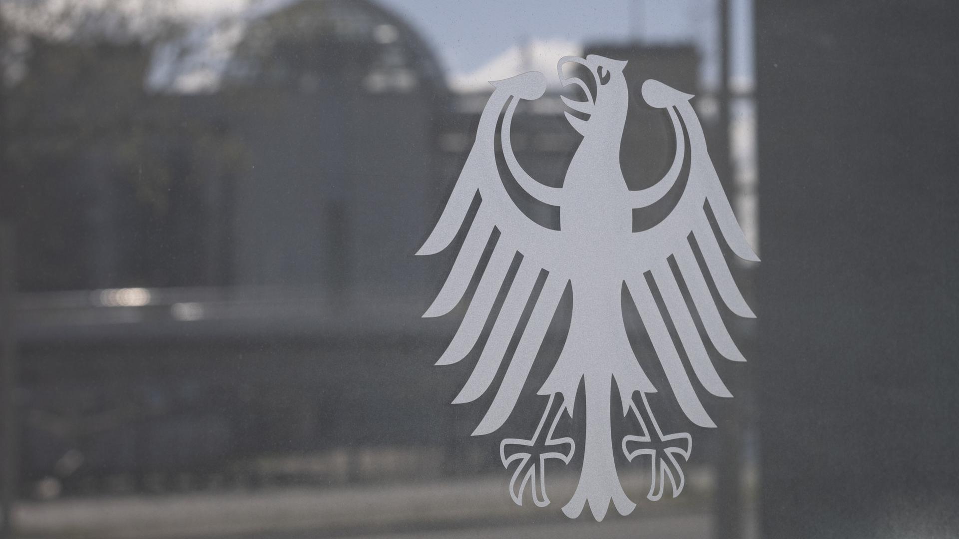 Der Bundesadler klebt an der Scheibe eines Ministeriums, in der Spiegelung des Glases sind der Bundestag und Reichstagskuppel sichtbar. 