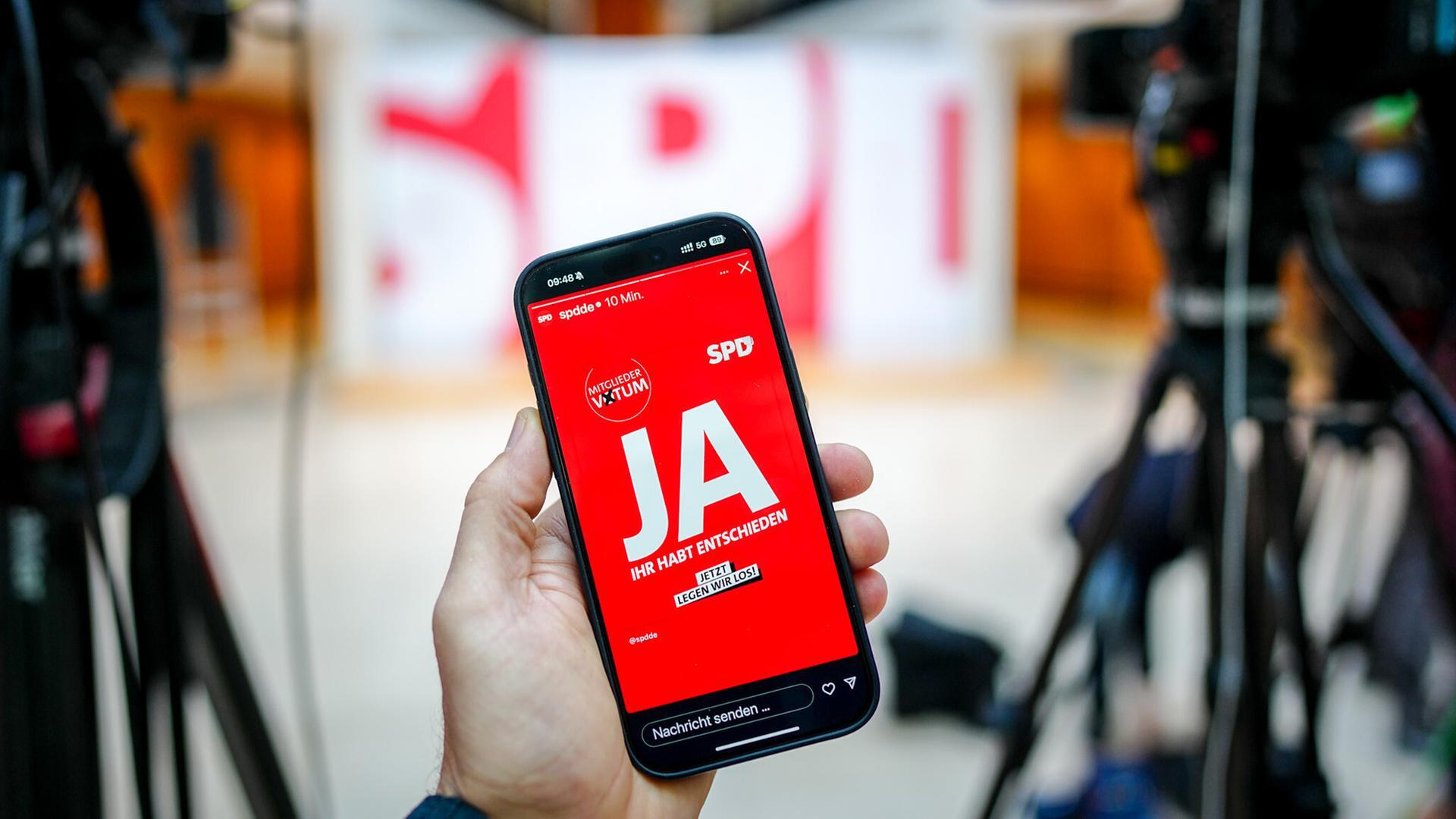 Auf einem Handy-Bildschirm ist auf einem roten Hintergrund das Wort „Ja“ zu lesen. Im Hintergrund sieht man in der Unschärfe einige aufgebaute Kameras für eine Pressekonferenz in der SPD-Parteizentrale. 