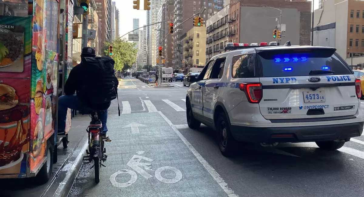 A photo of a biker waiting next to a police car at a traffic light.