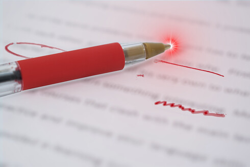 A red pen laying atop an edited manuscript with bold red editorial marks. The tip of the pen has a supernova flare.