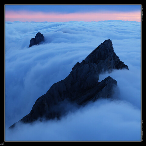 Mer de nuages bleutée et tourmentée, avec 2 crêtes triangulaires de sommets qui émergent. Une bande rose dans le ciel en haut de l'image