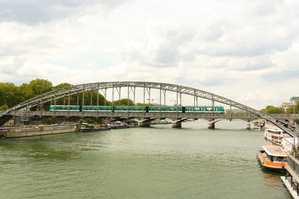 La Seine, enjambée par un long pont métallique. Le tablier et l'Arche du pont sont en structures métalliques. Une rame de métro, de type MF 01 (ici sur la ligne 5, mais qui circulent aussi sur les lignes 2 et 9) franchit le pont.