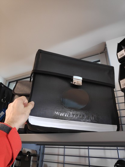 Détails d'une des sacoches de la photo précédente, prise de face.
Elle est noire avec une boucle de fermeture chromé au centre et une large bande blanche réfléchissante en bande.
Les reflets permettent de distinguer le logo de la Poste et l'URL abrégée de son site "www.laposte.fr" qui ont été masqués à l'encre noire.