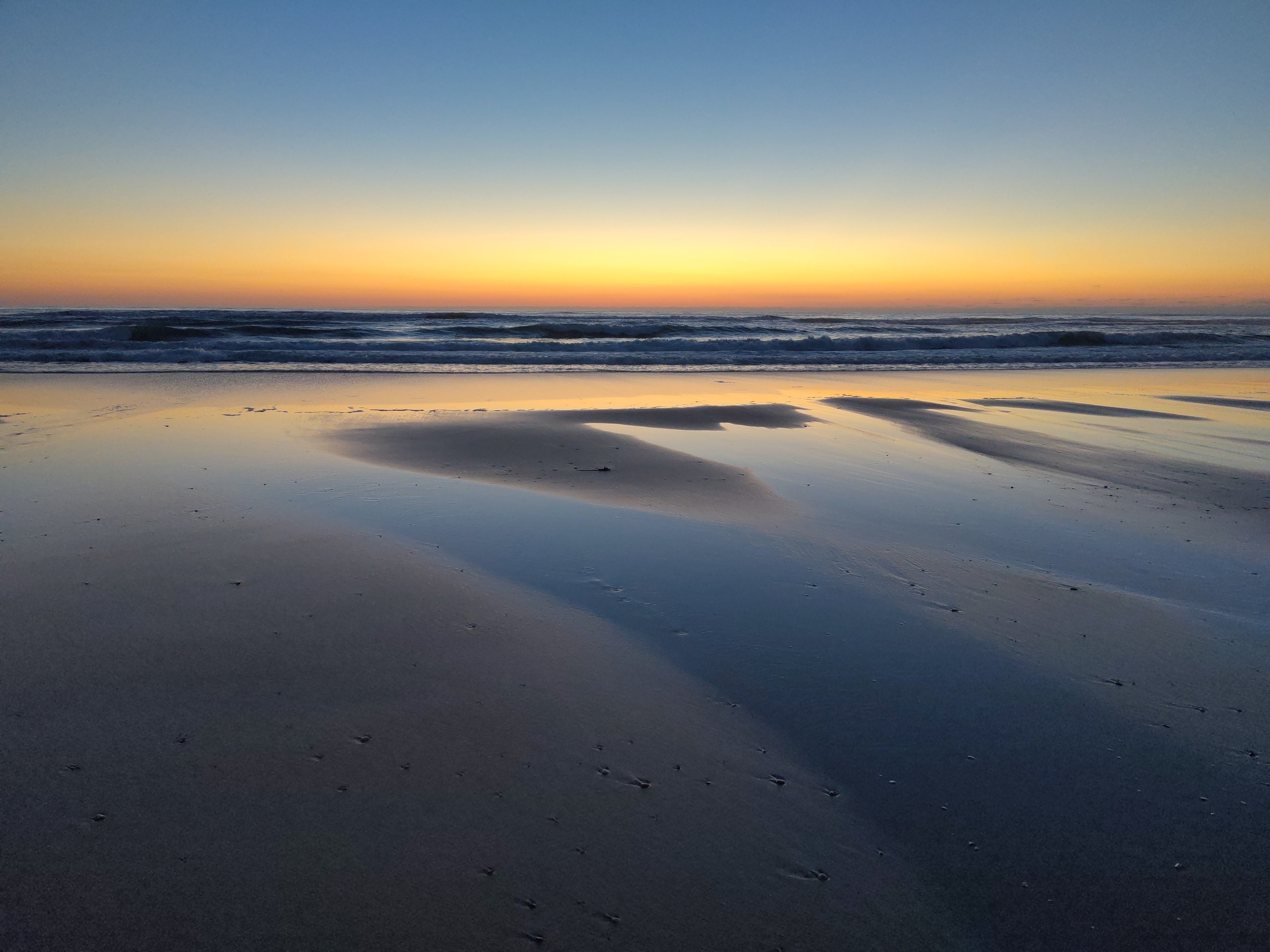 L'océan le soleil couché, couleurs orangées sur bleu et gris, un bout de plage et d'eau (marée descendante). Le calme des vagues.