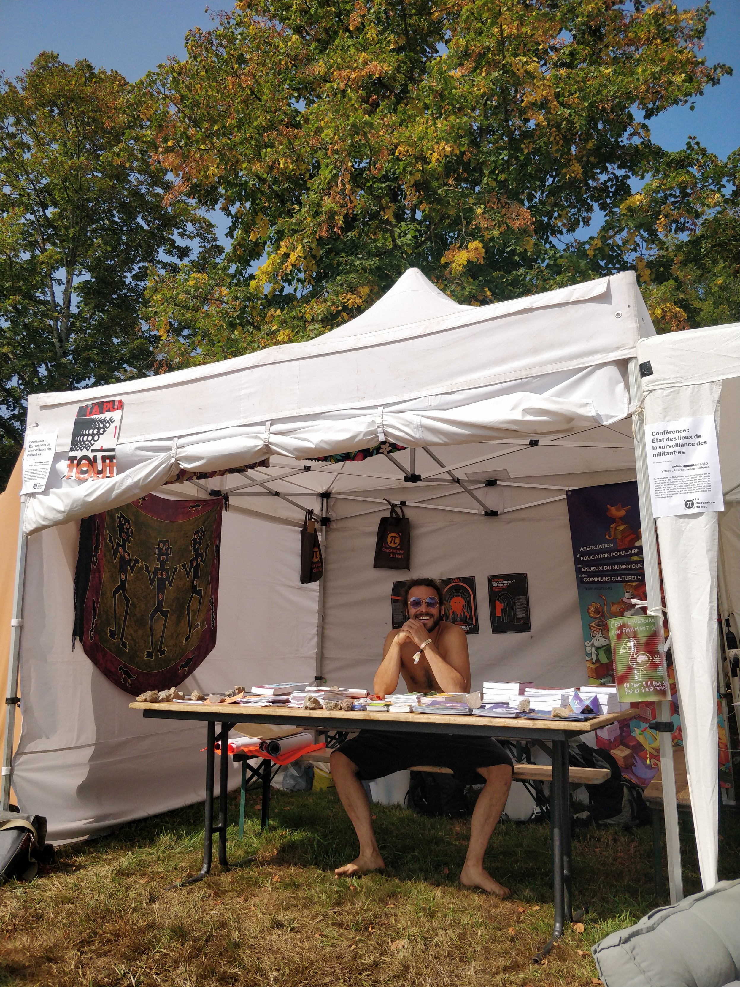 Un stand sur un festival avec des affiches et un humain derrière une table avec des brochures et des stickers. 