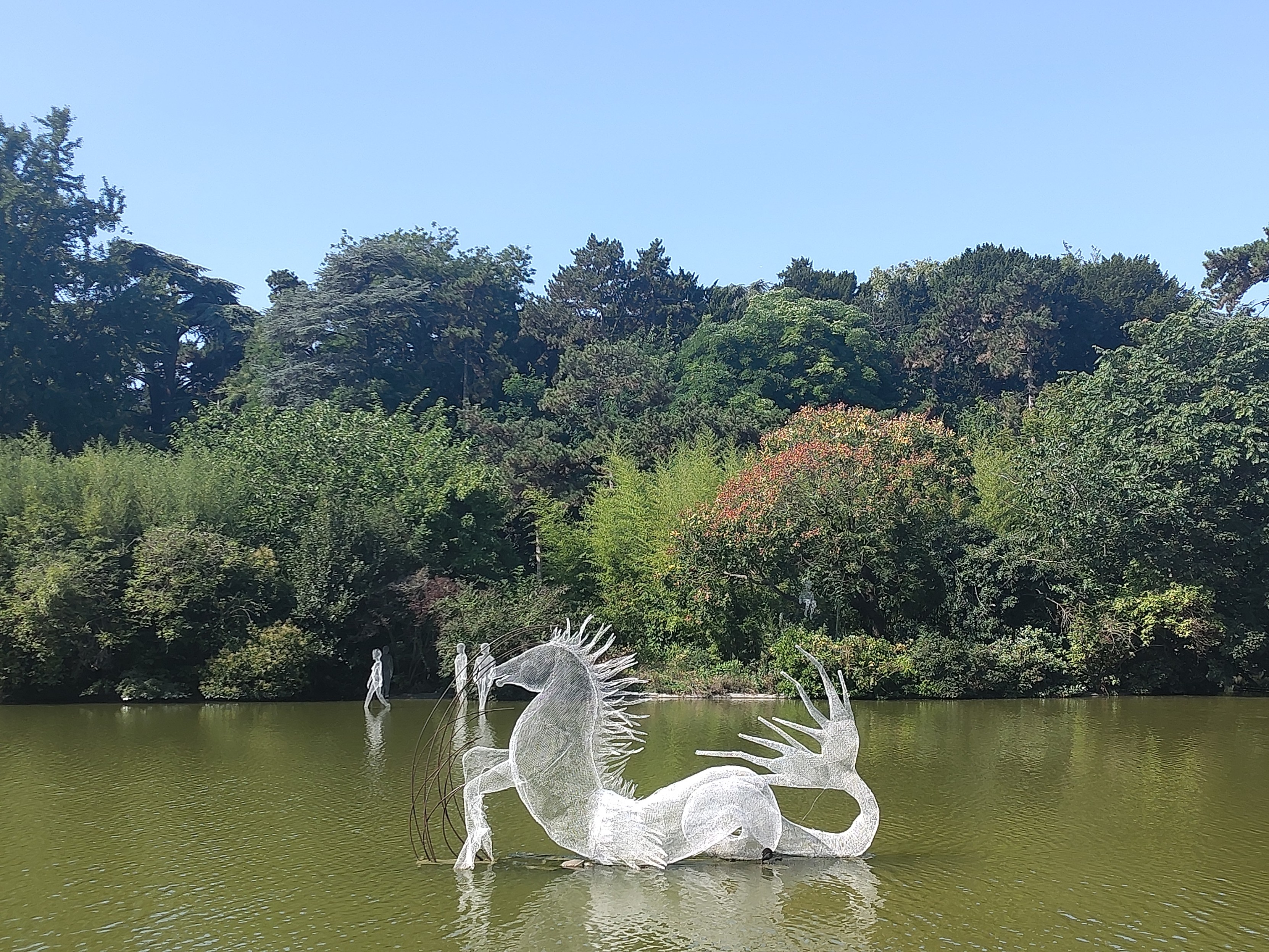 Un cheval sirène en grillage blanc métallique sur le plan d'eau. au fond, près des arbres, trois autres personnages plutôt enfantins.