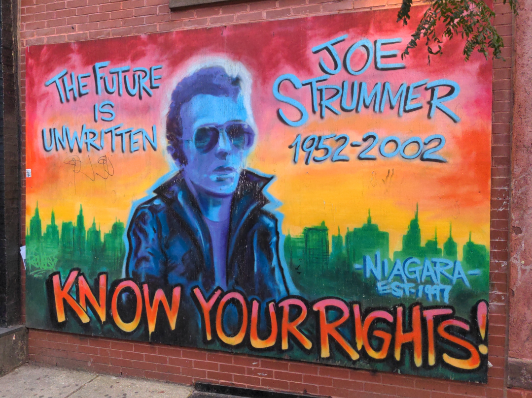 Sur une plaque couvrant un mur de briques, une fresque représentant un homme blanc de trois quarts, aux cheveux courts, portant lunette noire et veste en cuir, devant une skyline verte sur ciel dégradé de rouge à jaune, légendée "Joe Strummer <br />1952-2002&quot;, commentée &quot;The future is unwritten&quot; et &quot;Know your rights!&quot;, et signée &quot; Niagara est. 1997&quot;.