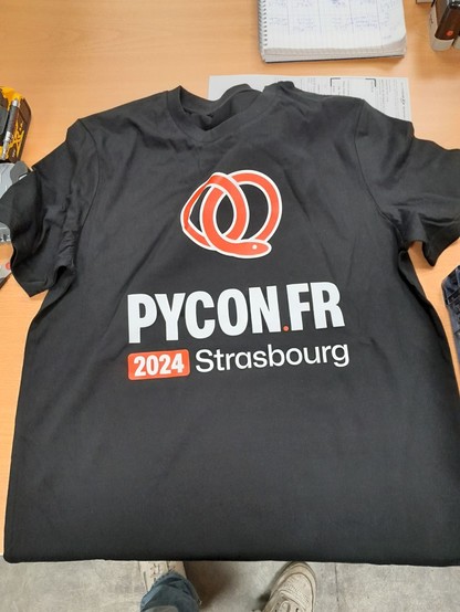 t-shirt pour la PyConFR 2024 à Strasbourg, t-shirt noir avec un logo sur le haut représentant un serpent rouge enroulé comme un bretzel, en-dessous se trouve les informations "PyConFR - 2024 Strasbourg"