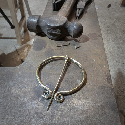 Une grande fibule en laiton (8cm de diamètre) avec ses deux extrémités spiralées, posée sur la table d'une grosse enclume, on voit quelques outils en haut du cadre (marteau, pinces) 