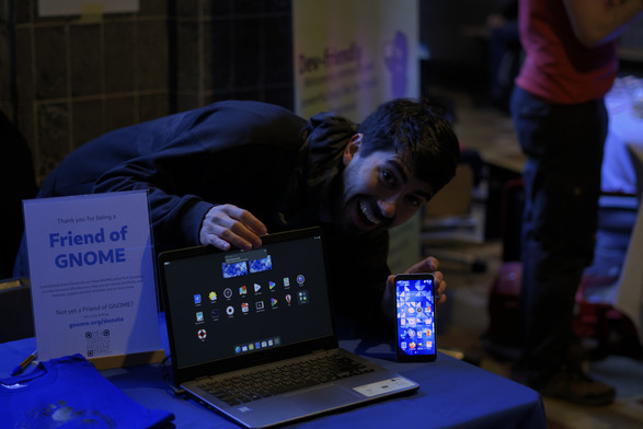 A man is smiling widely, while hunching on a laptop and a phone both running GNOME.

On the desk where the devices stand, there's a page saying "Friend of GNOME" and a pile of GNOME t-shirts.