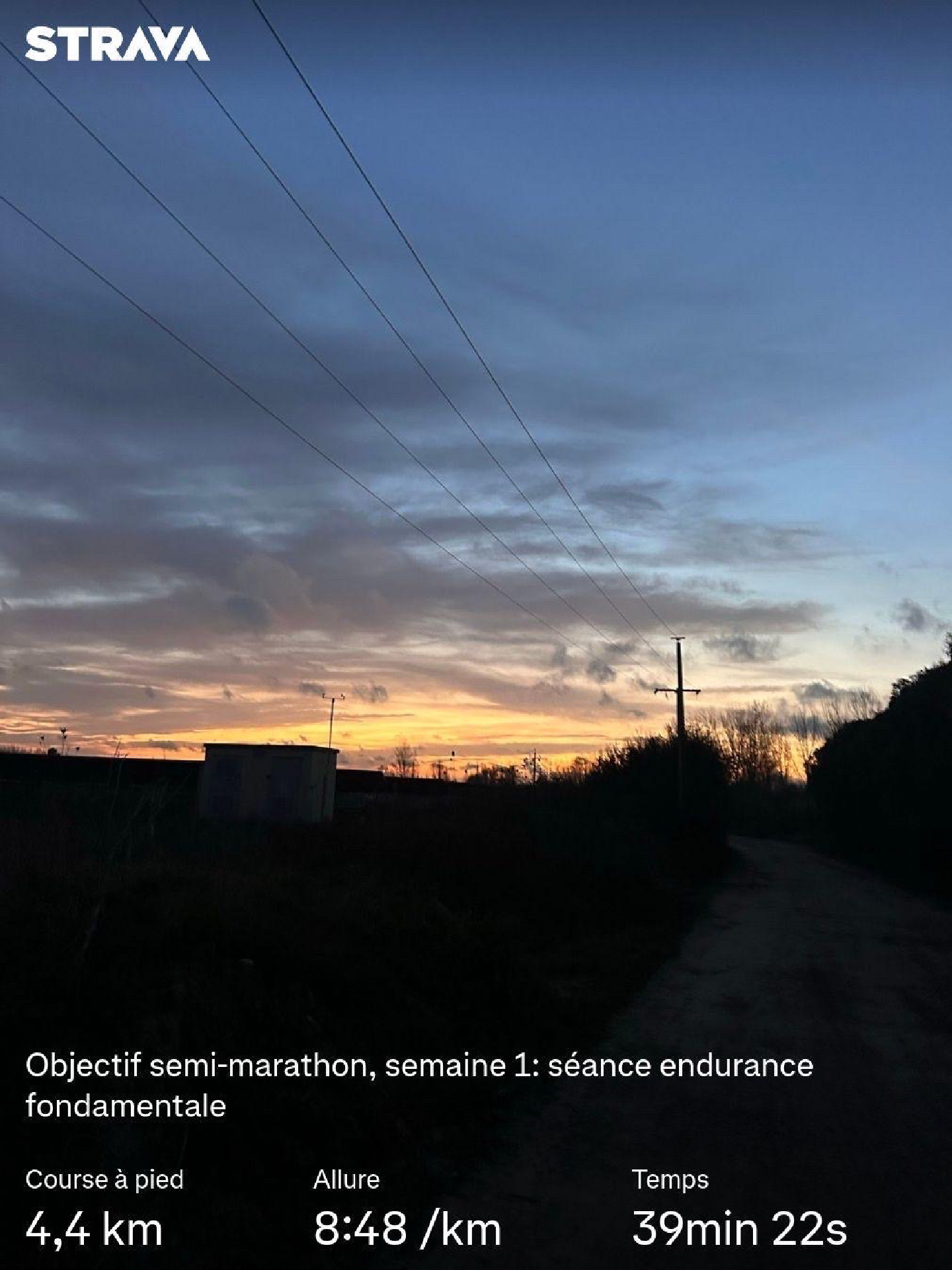 Image en contre-jour montrant le ciel clairsemé de nuages enflammé par le soleil levant. On devine des bâtiments industriels le long d’un chemin de terre. Des statistiques Strava sont affichées.