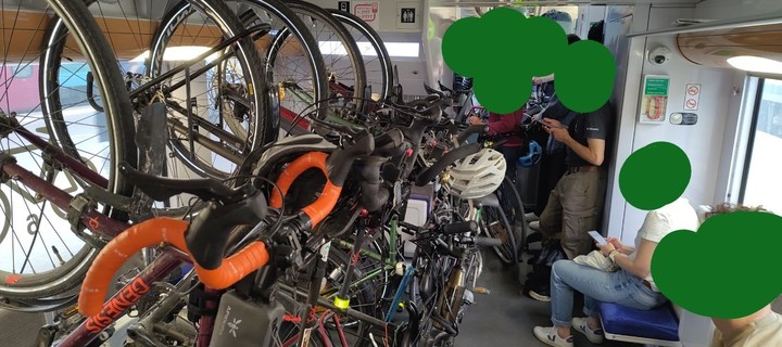 Une photo de cinq vélos accrochés au plafond, et de trois vélos posés devant et deux autres vélos au milieu des gens dans l'entrée du wagon de train