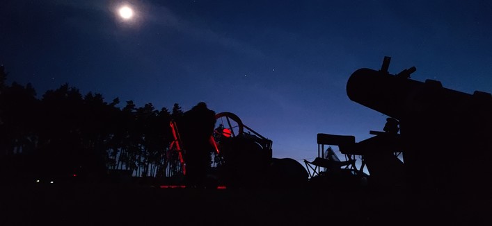 Crépuscule astronomique au-dessus du terrain d'observation. Les télescopes sont pointés sur la lune, ils apparaissent en ombres chinoises. Un observateur se tient aux-côtés de son télescope. Il porte une lampe frontale rouge.