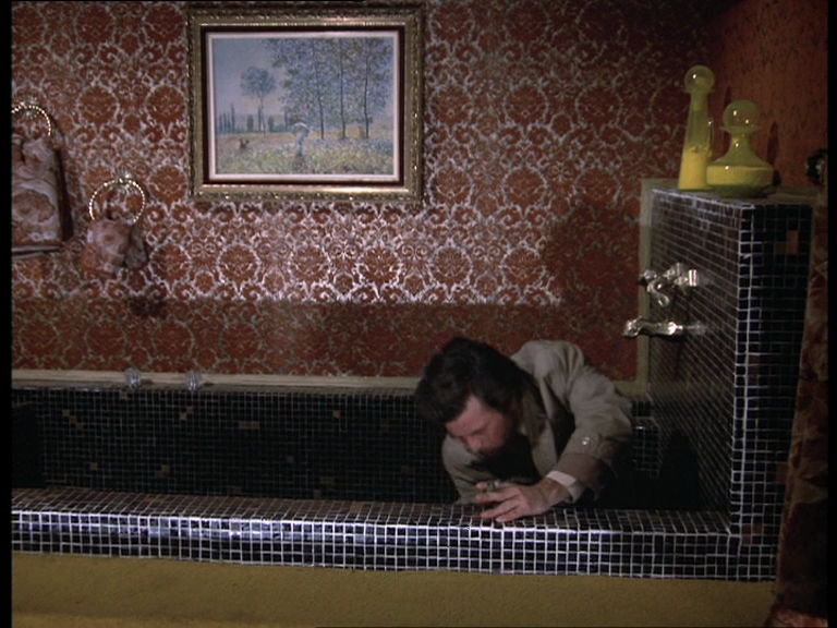 Columbo en train de fouiner accroupi dans une baignoire. La baignoire est en mosaïque bleue marine, avec des robinets en hauteur, deux gros flacons jaunes vifs posés sur le rebord. Le mur est couvert d'un incroyable papier peint rouge à motifs chamarrés, et au dessus de la baignoire est suspendu un tableau, une sorte d'aquarelle impressionniste.