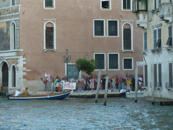 Gruppo di persone con cartelli "cessate il fuoco", "Gaza arriviamo", "Gaza we are coming" e bandiere palestinesi e della pace, al campo s. Zio a Venezia. Foto dal vaporetto per la stazione.