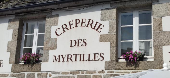 Devanture d'une crêperie, "crêperie des myrtilles". Des pots de fleurs roses posées sur les deux fenêtres entourant l'inscription.