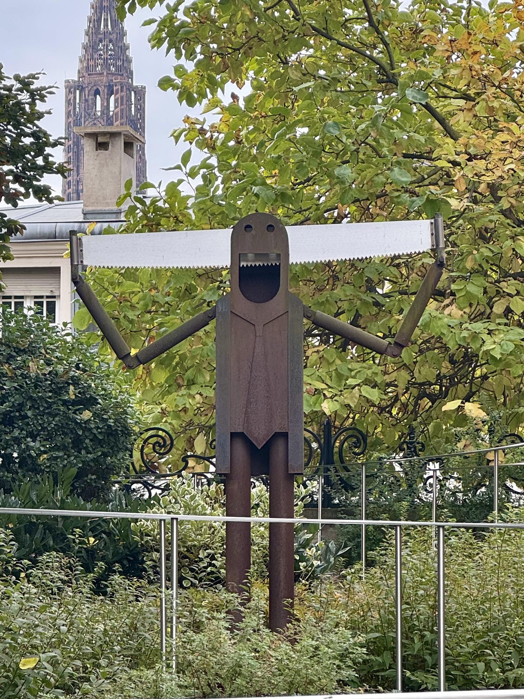 A photo in a public garden in Strasbourg. There’s a man made of metal with a saw going through his head. As he opens his mouth in a smile, the teeth of the saw look like his own teeth. In the background we can see the Strasbourg cathedral.