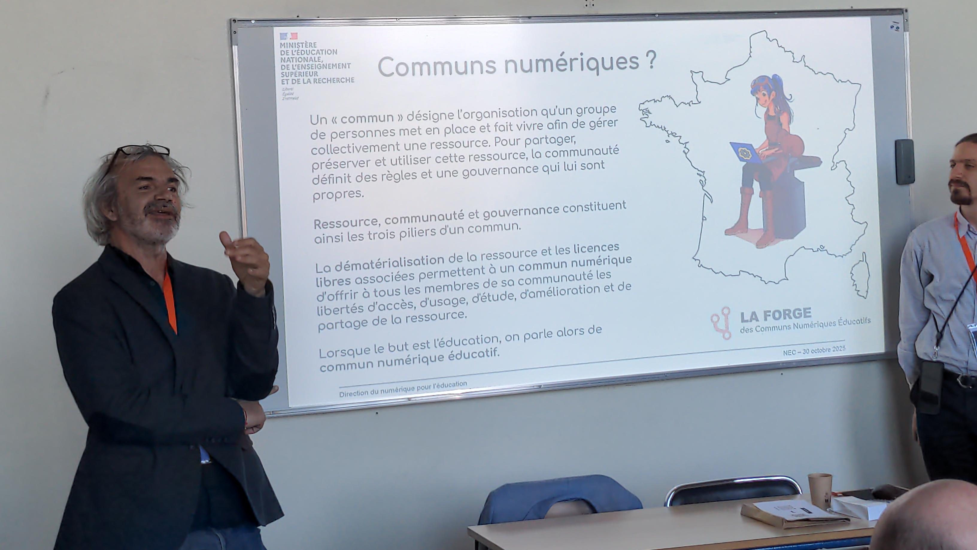 Alexis Kauffmann , debout à gauche d'un tableau, explique le contenu de la diapositive projetée au tableau, intitulée « Communs numériques »
Jean-Baptiste Clad est debout à droite du tableau.